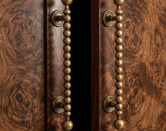 May include: Close-up of a wooden cabinet with two brass beaded handles. The cabinet doors are a dark brown with a unique wood grain pattern. The handles are ornate, with a large ball at the top and bottom.