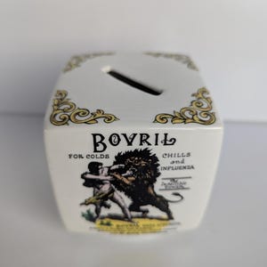 May include: A white ceramic Bovril money box with a slot on top and gold filigree corner designs. The front features a graphic of a man fighting a lion, with text that reads "Bovril for Colds, Chills and Influenza."