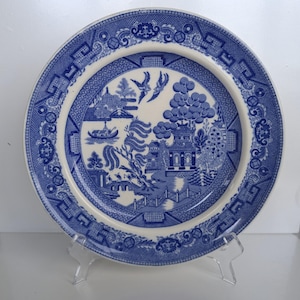 May include: Blue and white ceramic plate with a Willow pattern design. The plate features a central scene with a pagoda, bridge, boat, and birds. The rim has a decorative border. The plate is displayed on a clear stand.