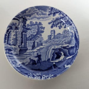 May include: A blue and white ceramic plate with a detailed landscape scene. The design features buildings, trees, and figures, all in a classic, vintage style. The plate has a slightly raised edge, typical of traditional tableware.