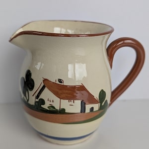 May include: Cream ceramic jug with a brown handle and rim. The jug has a hand-painted scene of a house with a brown roof, green trees, and a brown and blue stripe around the base. A charming addition to any kitchen.