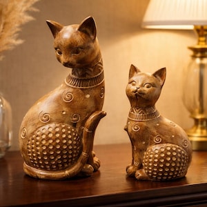 Könnte beinhalten: Zwei braune Katzenfiguren mit dekorativen Halsbändern und detaillierten Mustern. Die größere Katze sitzt und blickt nach unten, während die kleinere Katze aufrecht sitzt. Die Figuren stehen auf einer dunklen Holzoberfläche, was auf ein Wohnkulturthema hindeutet.