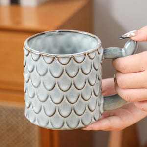 May include: A ceramic mug with a scale pattern in grey and brown. The handle is shaped like a tail. The mug is designed for hot drinks and is suitable for home or office use.