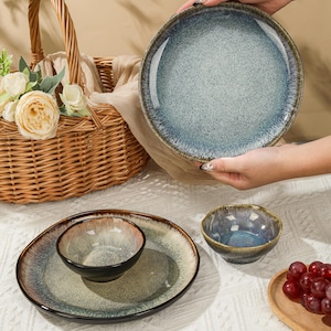 Könnte beinhalten: Ein Set aus Keramikgeschirr mit einer gesprenkelten blau-braunen Glasur. Das Set umfasst große Teller, kleine Schalen und eine Servierschüssel. Ein Weidenkorb mit Blumen befindet sich im Hintergrund.
