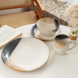 May include: A ceramic tableware set featuring a large plate, a bowl, a saucer, and a mug. The pieces have a white base with sections of black, tan, and brown. The set is arranged on a white tablecloth.