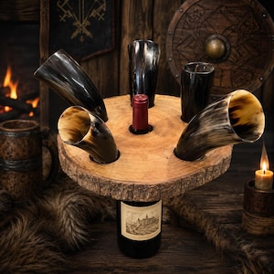 Pode incluir: Uma tábua circular de madeira com bordas de casca serve como suporte para quatro chifres de beber e uma garrafa de vinho central. Os chifres são escuros, polidos e curvos, enquanto a garrafa de vinho tem um rótulo creme com um castelo.
