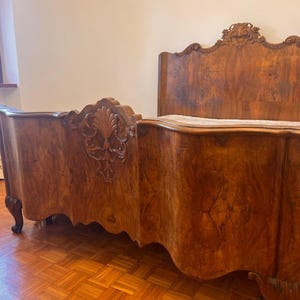 Puede incluir: Una estructura de cama de madera grande con un cabecero alto y un piecero con intrincados motivos tallados de conchas y volutas. La madera muestra un patrón de veta rico y arremolinado en tonos marrón medio con paneles curvos y ondulados.