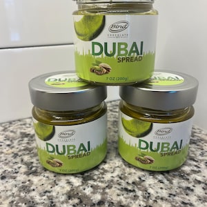 Puede incluir: Tres frascos de vidrio de Dubai Spread, cada uno con tapa plateada y etiqueta blanca. La etiqueta presenta el nombre de la marca y detalles del producto, incluyendo el texto "DUBAI SPREAD" y un gráfico de pistachos. Cada frasco contiene 200g de crema.