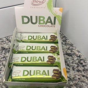 May include: A display box of "Dubai Chocolate" bars. The bars are in green and white wrappers with the text "Dubai" and a picture of chocolate. The box has the "Bind Chocolate" logo and the word "Dubai" in large letters.