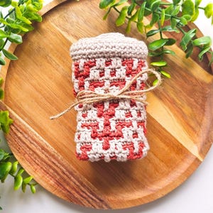 Könnte beinhalten: Eine handgefertigte Häkelhülle in Rot und Creme, mit Bindfaden gebunden, liegt auf einem Holztablett. Die Hülle hat ein geometrisches Muster und einen passenden Deckel. Grünzeug umgibt das Tablett.