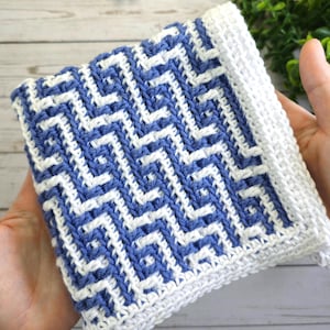 May include: A square, hand-crocheted washcloth with a geometric pattern in blue and white. The edges are finished with a solid white border. The washcloth is held in two hands.