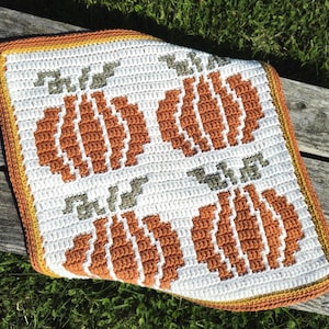 Op de afbeelding: Een gehaakte vierkante deken met een pompoenontwerp. De deken heeft een witte achtergrond met oranje en witte pompoenen en groene stelen. De rand is een mix van oranje, geel en bruin.