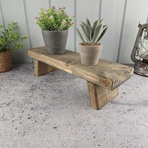 Könnte beinhalten: Ein rechteckiger Holzständer mit dunkelbraunem Finish und vier kurzen Beinen. Auf dem Ständer stehen ein grauer, gesprenkelter Topf mit einer grünen Pflanze mit kleinen rosa Blüten und ein kleinerer grauer Topf mit einer Sukkulente mit dicken Blättern.