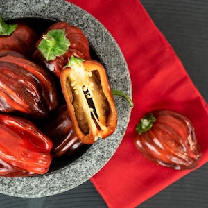 May include: A close-up of a gray stone bowl filled with red and brown peppers, one cut in half. A red napkin is in the background. The peppers have green stems and a striped pattern.