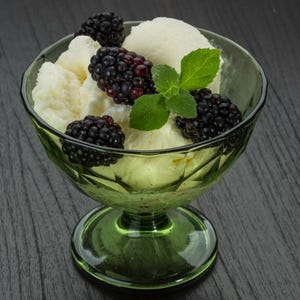 May include: A green glass dessert bowl filled with vanilla ice cream, fresh blackberries, and a sprig of mint. The ice cream is creamy white, and the blackberries are a deep purple. The bowl sits on a dark wooden surface.