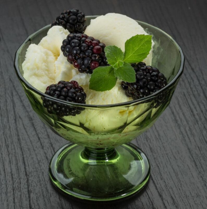 May include: A green glass dessert bowl filled with vanilla ice cream, fresh blackberries, and a sprig of mint. The ice cream is creamy white, and the blackberries are a deep purple. The bowl sits on a dark wooden surface.