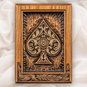 Op de afbeelding: Een rechthoekige houten plaat met een diep uitgesneden schoppensymbool in het midden. De schoppen is versierd met complexe bladerpatronen en een rond medaillon. De rand bevat uitgesneden veermotieven en een getextureerde houtnerf.