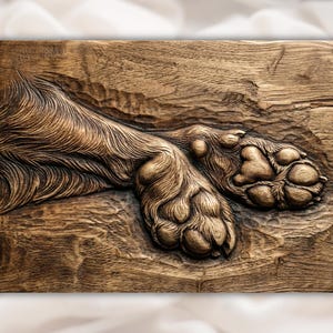 May include: A detailed, carved wooden relief depicting a dog's paw pads and fur. The artwork is rendered in shades of brown, showcasing the texture of the wood and the dog's fur. The relief is mounted on a rectangular wooden panel.