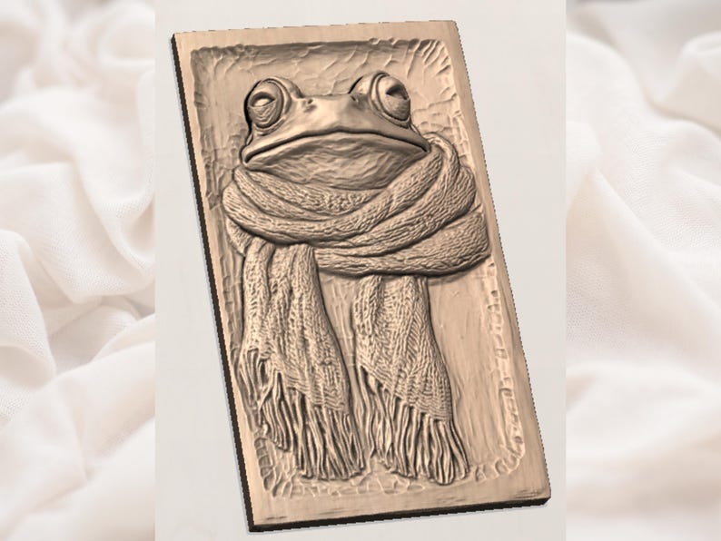 May include: A rectangular, brown relief carving of a frog wearing a scarf. The frog has large, round eyes and is centered on the rectangular plaque. The scarf is draped around the frog's neck and has a fringed edge.