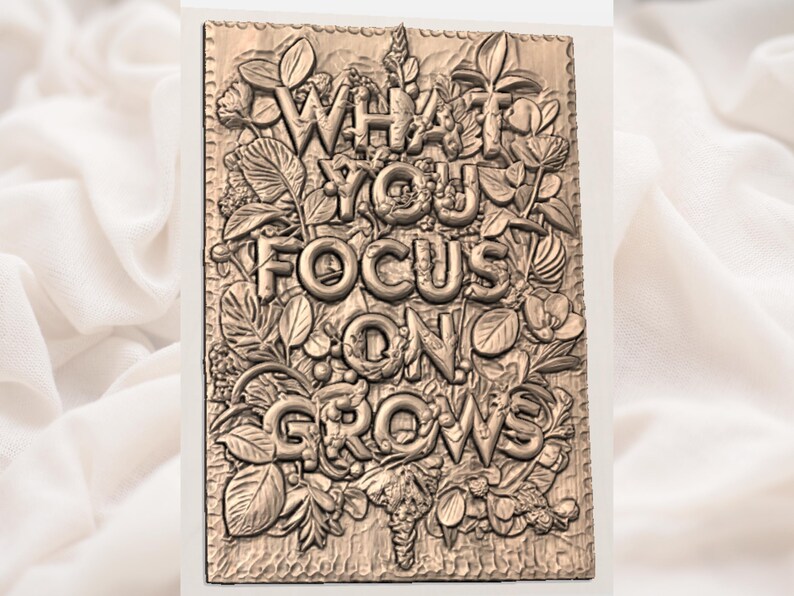 Puede incluir: Placa rectangular de color bronce con las palabras "WHAT YOU FOCUS ON GROWS" en letras en relieve y negritas. Las letras est&aacute;n rodeadas de elementos bot&aacute;nicos detallados, incluyendo hojas y flores, creando un efecto texturizado tridimensional.