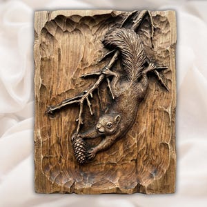 May include: A detailed wood carving of a squirrel hanging upside down from a branch, holding a pinecone. The carving is set against a textured wooden background, showcasing the natural grain and tones of the wood. The squirrel is brown and the wood is a warm brown.