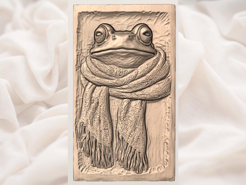 May include: A rectangular relief carving of a frog wearing a scarf. The frog has large, round eyes and a detailed scarf with fringe. The carving is in a neutral, earthy tone, suggesting a natural material.