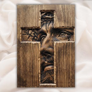 May include: Wooden wall art depicting a carved image of Jesus Christ within a cross-shaped cutout. The artwork shows detailed facial features, including a beard, eyes, and a crown of thorns. The wood has a warm, brown tone.