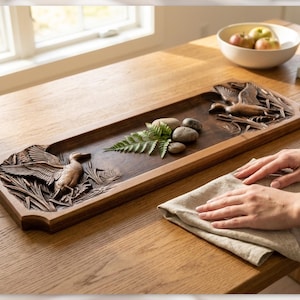 May include: A rectangular dark brown wooden tray featuring intricate carvings of ducks in flight among reeds at both ends. The center of the tray holds a small green fern frond and several smooth gray river stones arranged on the flat surface.