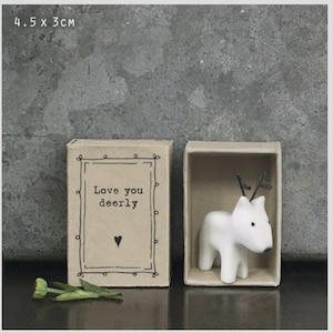 Puede incluir: Una pequeña figura de ciervo de cerámica blanca dentro de una caja tipo cerilla de 4,5 cm x 3 cm. La caja tiene el texto "Love you deerly" con un corazón. El ciervo tiene cuernos negros y una nariz negra.