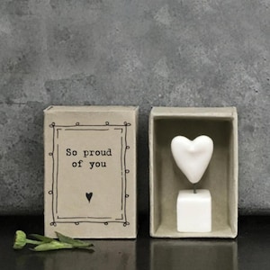 May include: A small, open cardboard box with the text "So proud of you" and a heart symbol next to a white heart-shaped ornament inside a similar box. A small green flower lies in front of the boxes.
