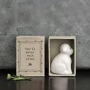 Puede incluir: Una figura de perro de cerámica blanca dentro de una caja, junto a una caja de cerillas con el texto "You'll never walk alone". La caja está abierta, revelando el perro. Un pequeño acento floral verde está en primer plano.