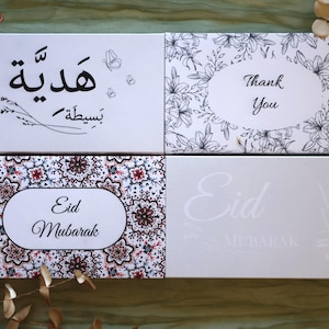 May include: Four white gift boxes with various designs. One box has Arabic script and butterflies, another says "Thank You" in a floral frame, and two others say "Eid Mubarak" with floral and script designs.