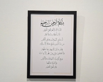 May include: A rectangular black frame containing a white print with black Arabic calligraphy. The text features the Bismillah followed by the Ayat al-Kursi, presented in a traditional script style with thin gold border lines on the white paper.