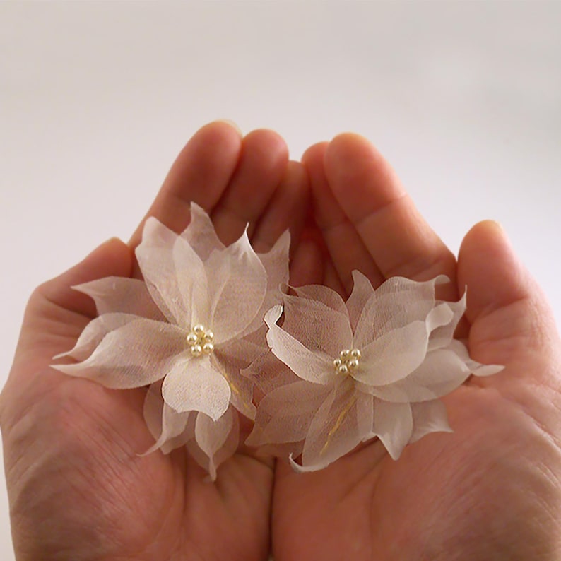K&ouml;nnte beinhalten: Zwei zarte wei&szlig;e Stoffblumen mit Perlenakzenten. Die Blumen werden in der Handfl&auml;che gehalten.