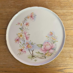 Op de afbeelding: Een witte keramische serveerschaal met een geschulpte rand, versierd met een bloemenontwerp. Het ontwerp toont roze, gele en perzik-kleurige bloemen met groene bladeren. De bloemen zijn gerangschikt in een delicaat, artistiek patroon.