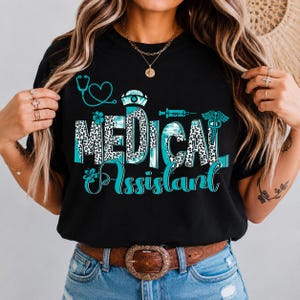 May include: Black t-shirt with the words "MEDICAL Assistant" in a teal and leopard print design. The design includes medical symbols like a stethoscope, syringe, and caduceus. The shirt has a crew neck and is worn with blue jeans and a brown belt.