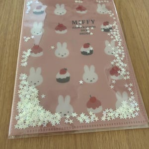 May include: A rectangular pink plastic folder featuring a repeating pattern of Miffy rabbit faces and small cakes with strawberries. The text MIFFY ILLUSTRATIONS DICK BRUNA is printed in the center. The edges contain loose white star-shaped sequins.