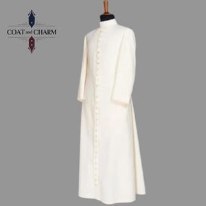 May include: Cream-colored, long-sleeved robe with a mandarin collar and a row of small buttons down the front. The robe is displayed on a mannequin. The logo "COAT and CHARM" is in the upper left corner.