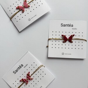 Può includere: Tre braccialetti a farfalla su biglietti bianchi. Ogni biglietto riporta le parole "Santéa" e "Studio". I braccialetti presentano disegni di farfalle in tonalità rosa e rosse, infilati su un cordino dorato.