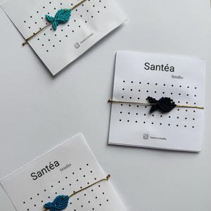Peut inclure: Trois bracelets poisson en macramé faits à la main sur des cartes blanches. Les poissons sont turquoise et noirs, attachés à des chaînes dorées. Chaque carte porte le texte "Santéa Studio" et un identifiant de réseau social.