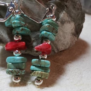 May include: Dangle earrings with turquoise and red coral beads, accented with silver-colored metal beads and lever-back closures. The earrings are displayed on a gray stone surface.
