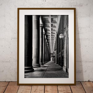 May include: Monochrome framed print showing a colonnade with tall columns and a long walkway. The image is in a brown frame and is displayed on a wooden floor against a white textured wall.