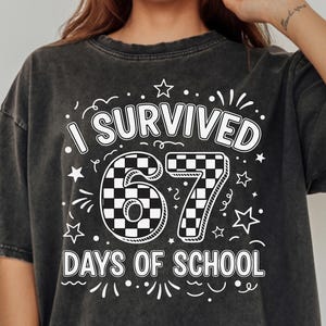 Peut inclure: T-shirt gris foncé avec le texte "I Survived 67 Days of School" en blanc. Le chiffre 67 a un motif à carreaux. Le t-shirt est de coupe ample.