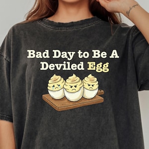 Può includere: T-shirt nero con la scritta "Bad Day to Be a Deviled Egg" e una grafica di tre uova alla diavola con facce scontrose su un tagliere di legno. La maglietta è di colore scuro e sbiadito.