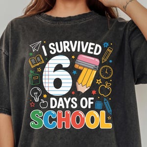 May include: Dark grey t-shirt with the text "I Survived 6 Days of School" in colourful letters. The graphic includes a pencil, apple, and other school-related icons. The shirt is a relaxed fit.