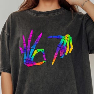 May include: Dark gray t-shirt with a rainbow skeleton hand graphic. The graphic shows a hand making the "OK" sign and another hand pointing down. The text "67" is also in the graphic. The shirt has a vintage, washed look.