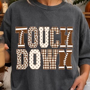 Peut inclure: Un t-shirt gris anthracite à manches courtes avec le texte TOUCH DOWN en grandes lettres capitales. Chaque lettre présente un motif unique, incluant des lacets de ballon de football, des taches de léopard, des pois et un motif à carreaux.