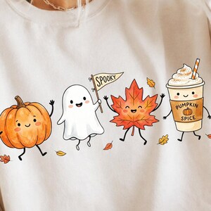 Peut inclure: Un sweat-shirt à col rond couleur crème avec une impression graphique représentant une citrouille orange souriante, un fantôme blanc tenant un drapeau marqué SPOOKY, une feuille d'érable rouge et un café épicé à la citrouille avec une paille.