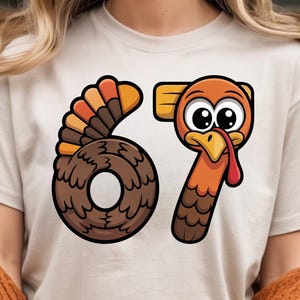Peut inclure: T-shirt beige avec un motif de dinde de dessin animé et le chiffre 67. Le chiffre 6 représente le corps de la dinde, et le chiffre 7 est sa tête et son cou. La dinde est orange, marron et jaune.