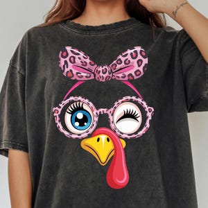 Peut inclure: T-shirt gris foncé surdimensionné avec un motif de visage de dinde de dessin animé. La dinde porte des lunettes, un nœud papillon et un œil qui cligne de l'œil. Le bec est jaune et le barbillon est rouge.